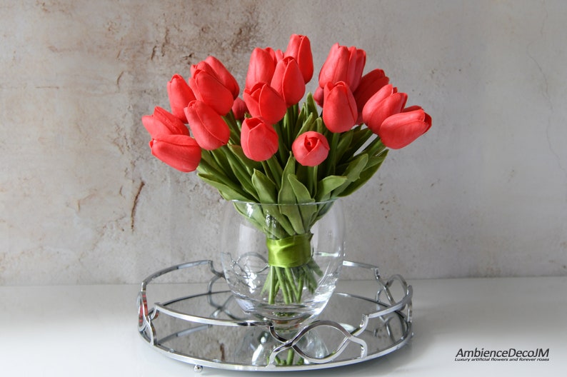 May include: A bouquet of red tulips arranged in a clear glass vase with a green ribbon. The vase is sitting on a silver tray with a mirrored surface.