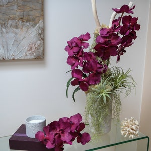 Könnte beinhalten: Ein Glastisch mit einer Vase mit lila Orchideen und grünen Pflanzen. Ein weißes Korallenstück und eine kleine graue Kerze stehen ebenfalls auf dem Tisch.