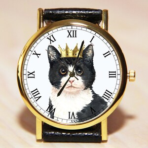 Op de afbeelding: Een gouden polshorloge met een zwarte leren band. De wijzerplaat van het horloge heeft een zwart-witte kat met een gouden kroon. De wijzerplaat met Romeinse cijfers is wit.