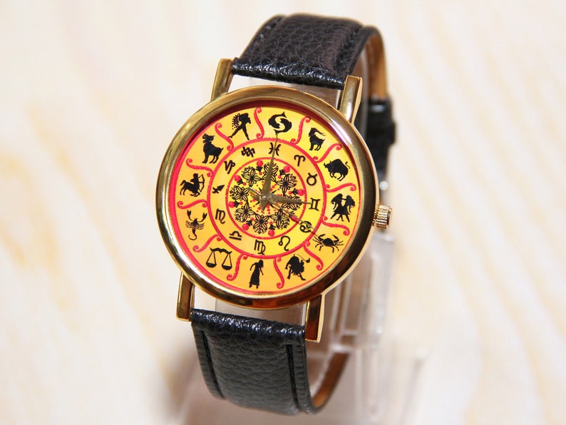 May include: A gold-tone wristwatch with a black leather band. The watch face features a yellow background with a red circle in the center and black zodiac symbols around the outer edge.