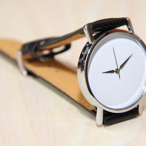 Pu&ograve; includere: Un orologio da polso argentato con quadrante bianco e cinturino in pelle nera. L'orologio ha un design semplice con due lancette e senza numeri.
