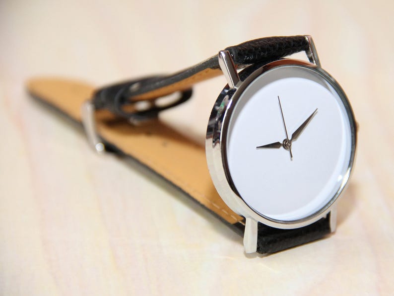 Pu&ograve; includere: Un orologio da polso argentato con quadrante bianco e cinturino in pelle nera. L'orologio ha un design semplice con due lancette e senza numeri.