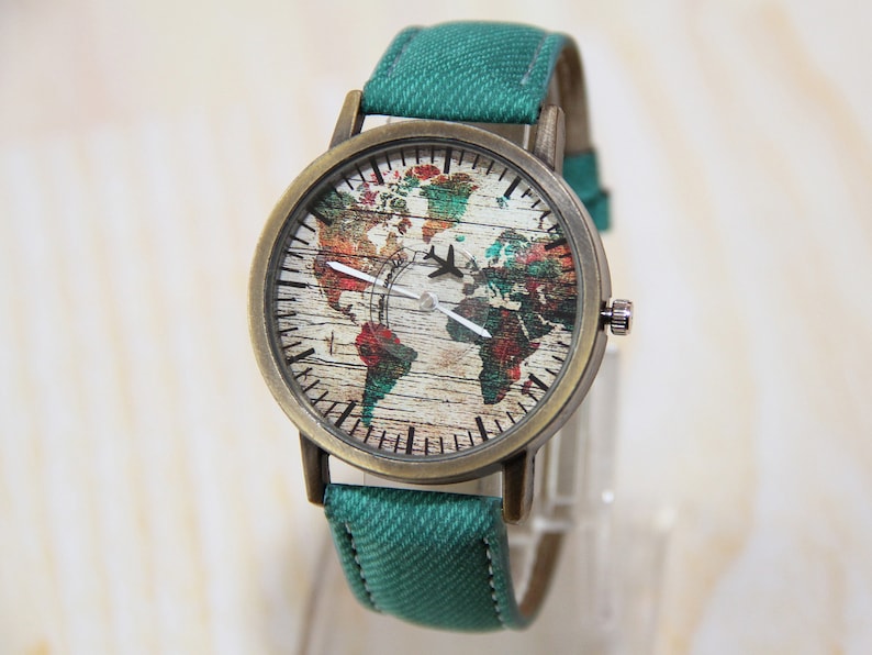 May include: A green canvas band watch with a round, bronze-colored case. The watch face features a vintage-style world map with a small airplane graphic.