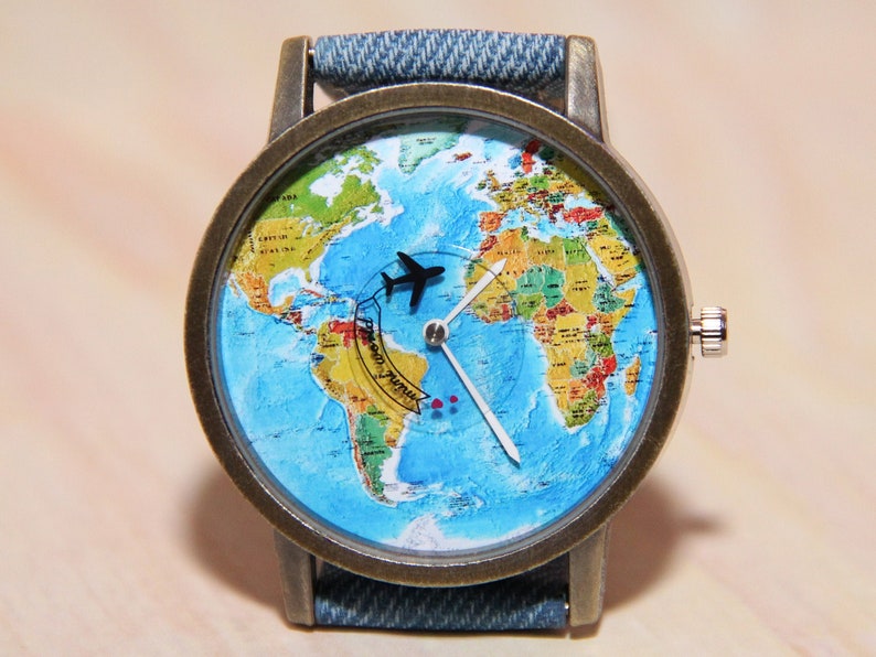 May include: A wristwatch with a denim strap and a round face featuring a world map with a small airplane icon. The watch face also has a white clock hand and a black clock hand.