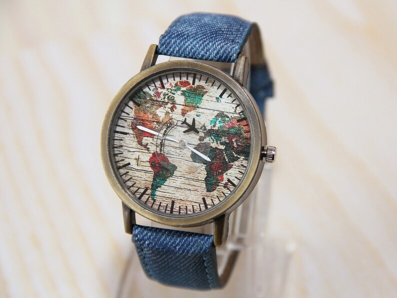 May include: A wristwatch with a denim band and a round face featuring a world map with a plane on it. The watch face is a vintage style with a wood grain background.