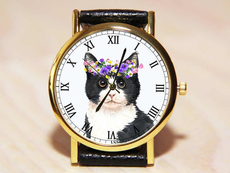 Op de afbeelding: Een gouden polshorloge met een zwarte leren band. De wijzerplaat van het horloge heeft een zwart-witte kat met een bloemenkrans. Romeinse cijfers markeren de uren.