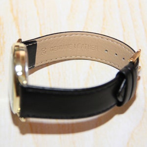 Puede incluir: Un reloj dorado con una correa de cuero negro. La correa est&aacute; etiquetada con "20 GENUINE LEATHER".