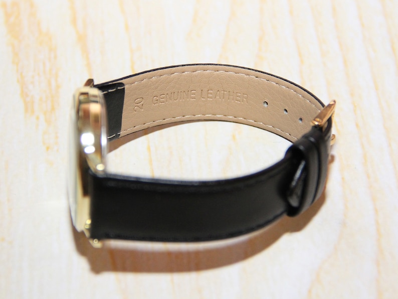 Puede incluir: Un reloj dorado con una correa de cuero negro. La correa est&aacute; etiquetada con "20 GENUINE LEATHER".