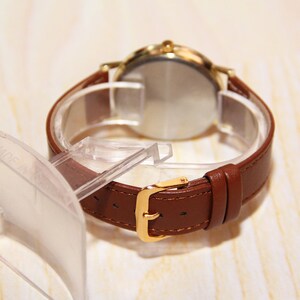 Puede incluir: Un reloj de pulsera de cuero marr&oacute;n con una caja dorada y una esfera transparente. El reloj est&aacute; en un expositor de pl&aacute;stico transparente.