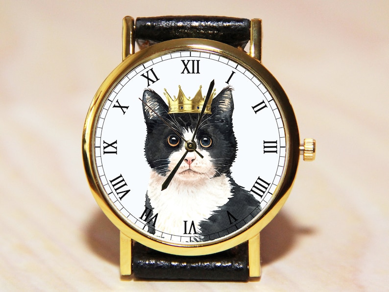 Op de afbeelding: Een gouden polshorloge met een zwarte leren band. De wijzerplaat van het horloge heeft een zwart-witte kat met een gouden kroon. De wijzerplaat met Romeinse cijfers is wit.