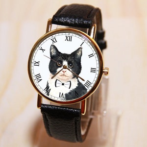 Op de afbeelding: Een gouden horloge met een zwarte leren band heeft een zwart-witte kat met een strikje op de wijzerplaat.