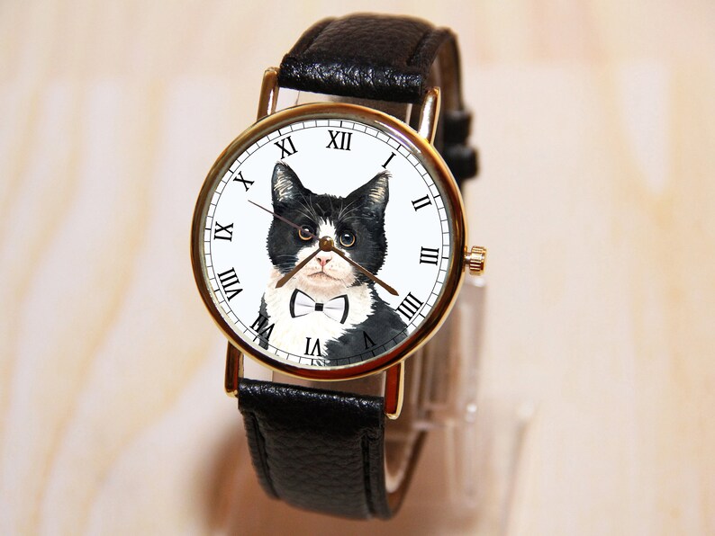 Op de afbeelding: Een gouden horloge met een zwarte leren band heeft een zwart-witte kat met een strikje op de wijzerplaat.