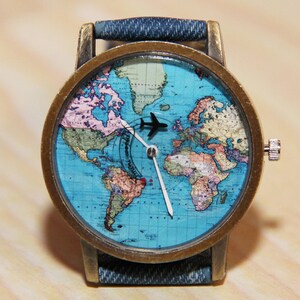 May include: A wristwatch with a blue denim strap and a round, brass-coloured case. The watch face features a world map with a small aeroplane icon in the centre.