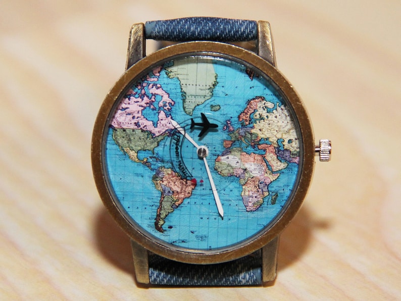 May include: A wristwatch with a blue denim strap and a round, brass-coloured case. The watch face features a world map with a small aeroplane icon in the centre.