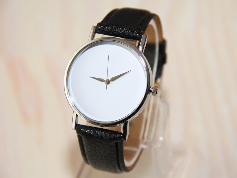 Pu&ograve; includere: Un orologio da polso minimalista e semplice con un quadrante bianco e lancette argentate. L'orologio ha un cinturino in pelle nera.
