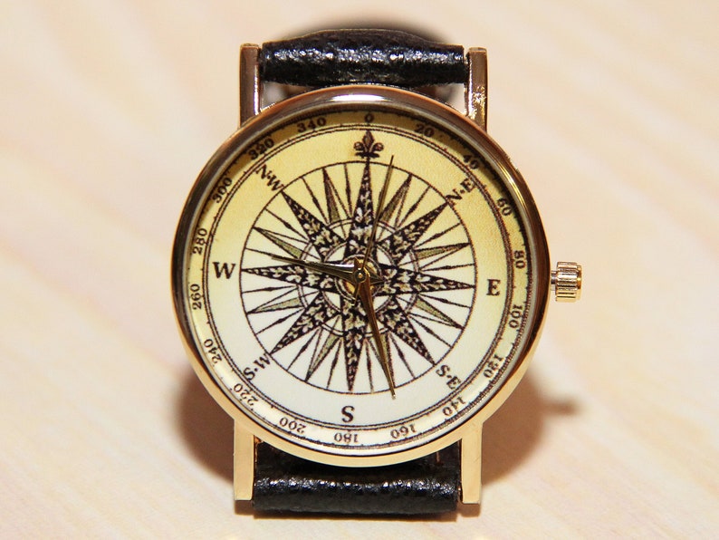 Puede incluir: Un reloj de pulsera dorado con una correa de cuero negro. La esfera del reloj presenta un dise&ntilde;o de br&uacute;jula con fondo blanco y marcas negras. Los puntos de la br&uacute;jula est&aacute;n etiquetados con las direcciones cardinales: N, E, S y W.