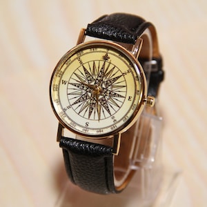 Puede incluir: Un reloj de pulsera dorado con una correa de cuero negro. La esfera del reloj presenta un dise&ntilde;o de br&uacute;jula con fondo blanco y marcas negras.