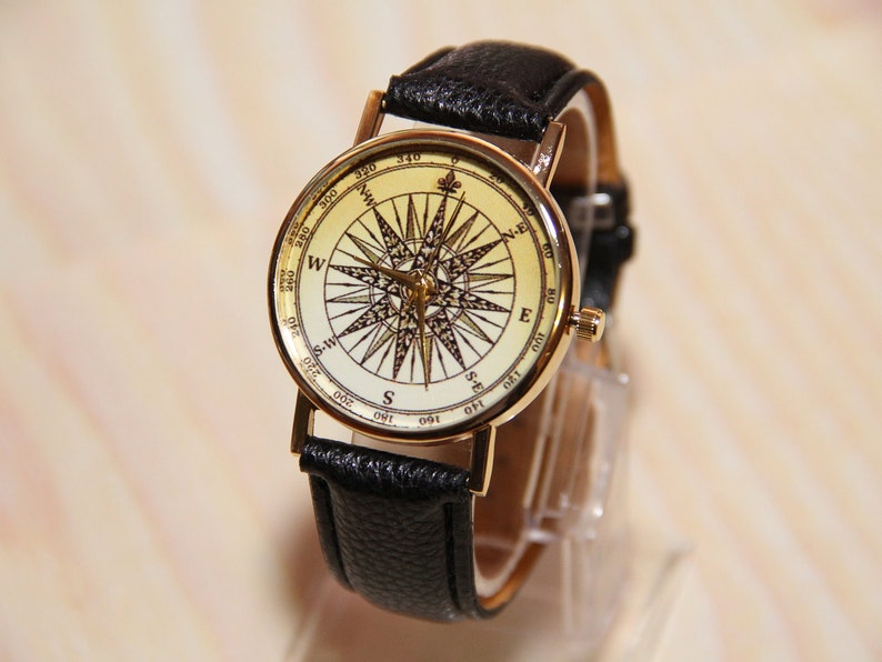 Puede incluir: Un reloj de pulsera dorado con una correa de cuero negro. La esfera del reloj presenta un dise&ntilde;o de br&uacute;jula con fondo blanco y marcas negras.
