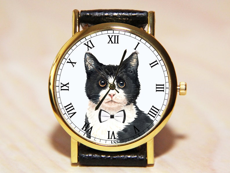 Op de afbeelding: Een gouden polshorloge met een zwarte leren band. De wijzerplaat van het horloge heeft een zwart-witte kat met een strikje en Romeinse cijfers voor de uren.