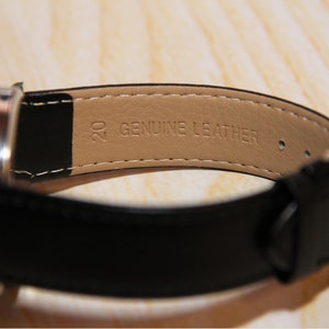 May include: A black leather watch band with a brown leather lining. The lining has the text "GENUINE LEATHER" printed on it.