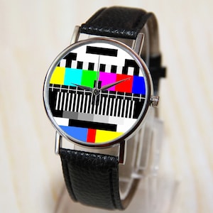 Op de afbeelding: Een polshorloge met een zwarte leren band, een zilveren kast en een kleurrijk televisie-testpatroon op de wijzerplaat.