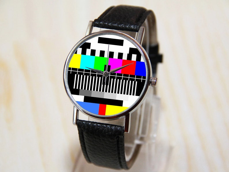 Op de afbeelding: Een polshorloge met een zwarte leren band, een zilveren kast en een kleurrijk televisie-testpatroon op de wijzerplaat.