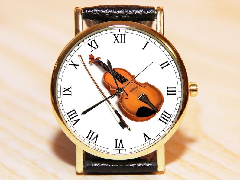 Peut inclure: Une montre-bracelet dor&eacute;e avec un cadran blanc et un bracelet en cuir noir. Le cadran pr&eacute;sente des chiffres romains pour les heures et un violon avec un archet.