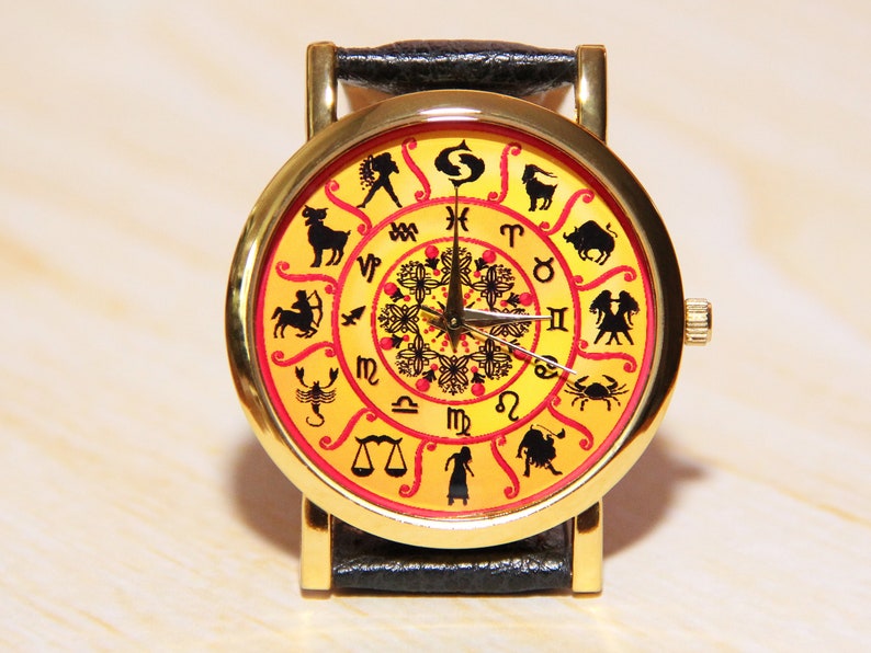 May include: A gold-tone wristwatch with a black leather band. The watch face features a yellow background with red accents and black zodiac symbols.