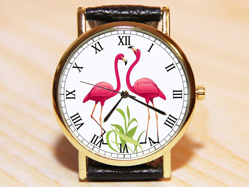 May include: A gold-tone wristwatch with a white face and black leather band. The face features a pair of pink flamingos and Roman numeral hour markers.