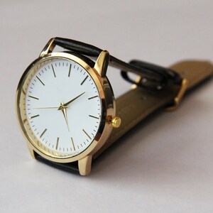 Puede incluir: Un reloj de pulsera dorado con esfera blanca y correa de cuero negra.