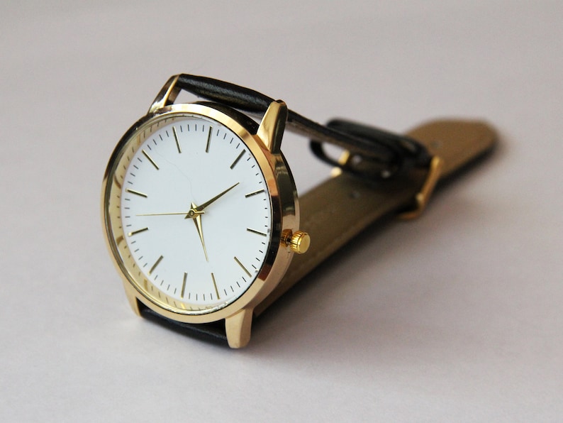 Puede incluir: Un reloj de pulsera dorado con esfera blanca y correa de cuero negra.