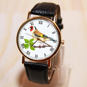 Peut inclure: Montre-bracelet dor&eacute;e avec un bracelet en cuir noir. Le cadran de la montre pr&eacute;sente une illustration d&eacute;taill&eacute;e d'un chardonneret perch&eacute; sur une branche avec des feuilles vertes. Chiffres romains.