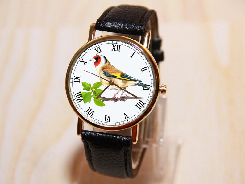 Peut inclure: Montre-bracelet dor&eacute;e avec un bracelet en cuir noir. Le cadran de la montre pr&eacute;sente une illustration d&eacute;taill&eacute;e d'un chardonneret perch&eacute; sur une branche avec des feuilles vertes. Chiffres romains.