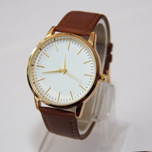 Puede incluir: Un reloj de pulsera dorado con esfera blanca y correa de cuero marr&oacute;n.
