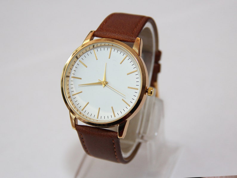 Puede incluir: Un reloj de pulsera dorado con esfera blanca y correa de cuero marr&oacute;n.