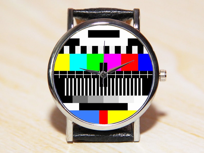 Op de afbeelding: Een polshorloge met een zwarte leren band en een zilveren wijzerplaat. De wijzerplaat van het horloge heeft een kleurrijk televisietestpatroon.