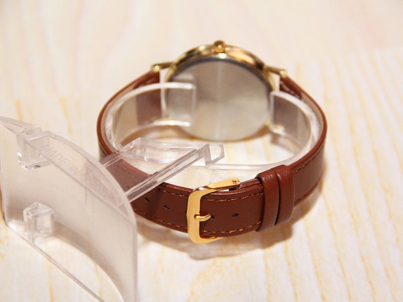 Peut inclure: Une montre &agrave; bracelet en cuir marron avec un bo&icirc;tier et une boucle dor&eacute;s. La montre est pr&eacute;sent&eacute;e sur un pr&eacute;sentoir en plastique transparent, montrant le dos du cadran. Le bracelet mesure environ 20 cm de long.