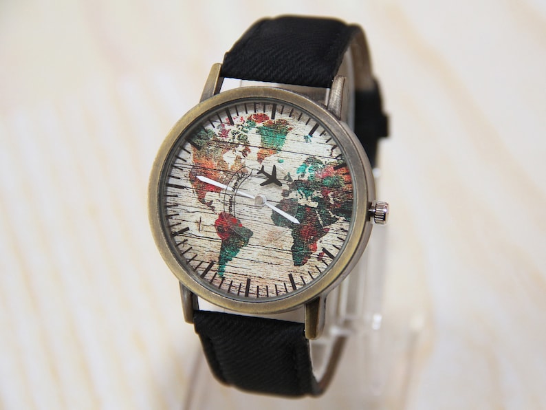 May include: A vintage-style wristwatch with a black canvas band and a round, brass-colored case. The watch face features a colorful world map with a woodgrain texture and a small airplane icon.