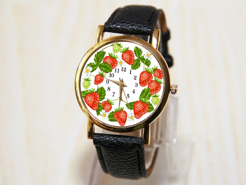 Op de afbeelding: Een gouden polshorloge met een witte wijzerplaat en een zwarte leren band. De wijzerplaat van het horloge heeft een ontwerp van rode aardbeien en groene bladeren.