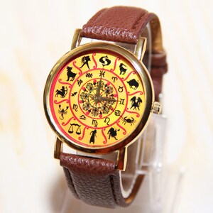 May include: A gold-tone wristwatch with a brown leather band. The watch face features a yellow background with a red circle in the center. The circle is decorated with a floral design and surrounded by the 12 zodiac signs.