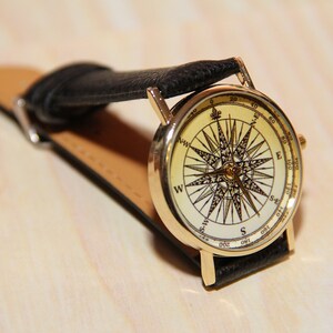Puede incluir: Un reloj de pulsera dorado con una correa de cuero negro. La esfera del reloj presenta un dise&ntilde;o de br&uacute;jula con fondo blanco y marcas negras.