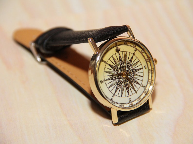 Puede incluir: Un reloj de pulsera dorado con una correa de cuero negro. La esfera del reloj presenta un dise&ntilde;o de br&uacute;jula con fondo blanco y marcas negras.