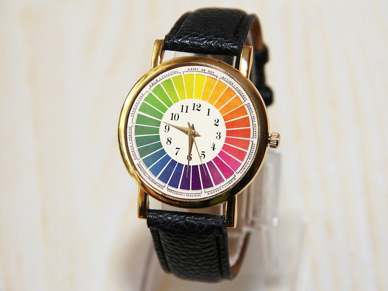 Puede incluir: Un reloj de pulsera dorado con una correa de cuero negro. La esfera del reloj presenta una rueda de colores arco&iacute;ris con los n&uacute;meros del 1 al 12 alrededor del borde.