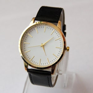 Puede incluir: Un reloj dorado con esfera blanca y correa de cuero negra.