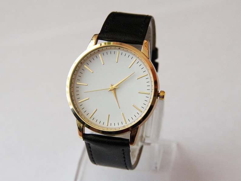 Puede incluir: Un reloj dorado con esfera blanca y correa de cuero negra.