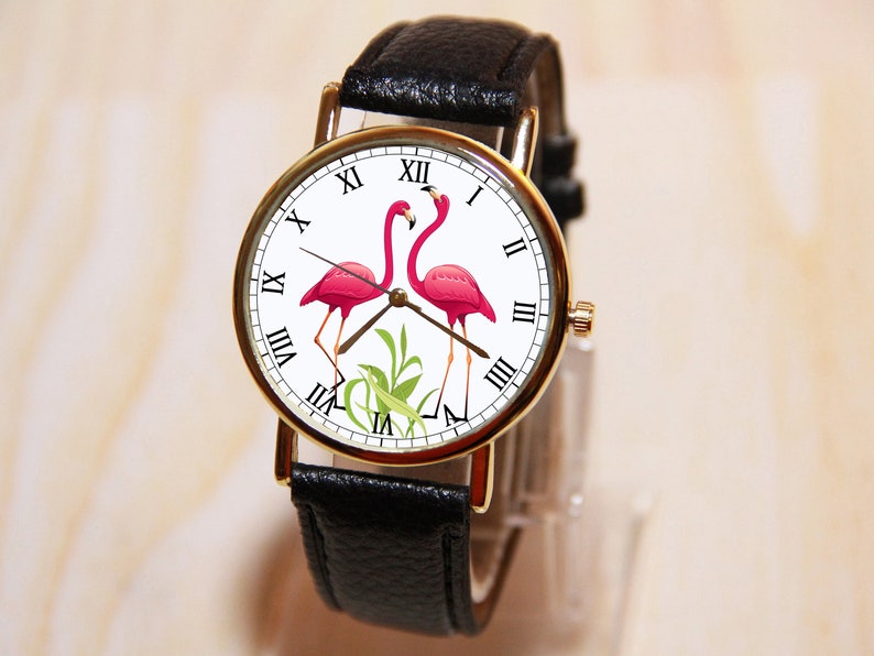 May include: A black leather wristwatch with a white face and gold accents. The face features two pink flamingos and Roman numeral hour markers.