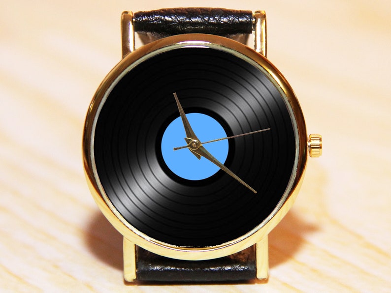 May include: A gold-tone wristwatch with a black leather band. The watch face features a black vinyl record with a blue center and gold-tone hands.