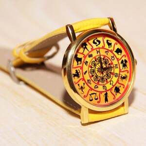 May include: A yellow wristwatch with a gold-colored face featuring the zodiac signs. The watch face has a red and yellow background with black zodiac symbols.