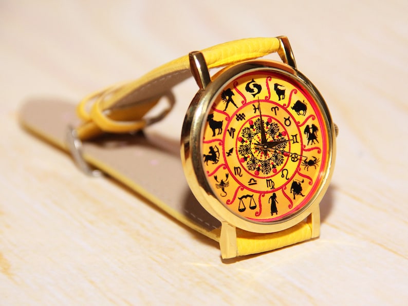 May include: A yellow wristwatch with a gold-colored face featuring the zodiac signs. The watch face has a red and yellow background with black zodiac symbols.