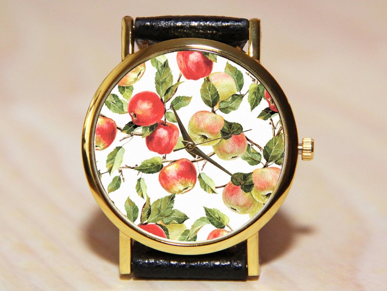 May include: A gold-tone wristwatch with a black leather band and a white face featuring a repeating pattern of red and green apples.